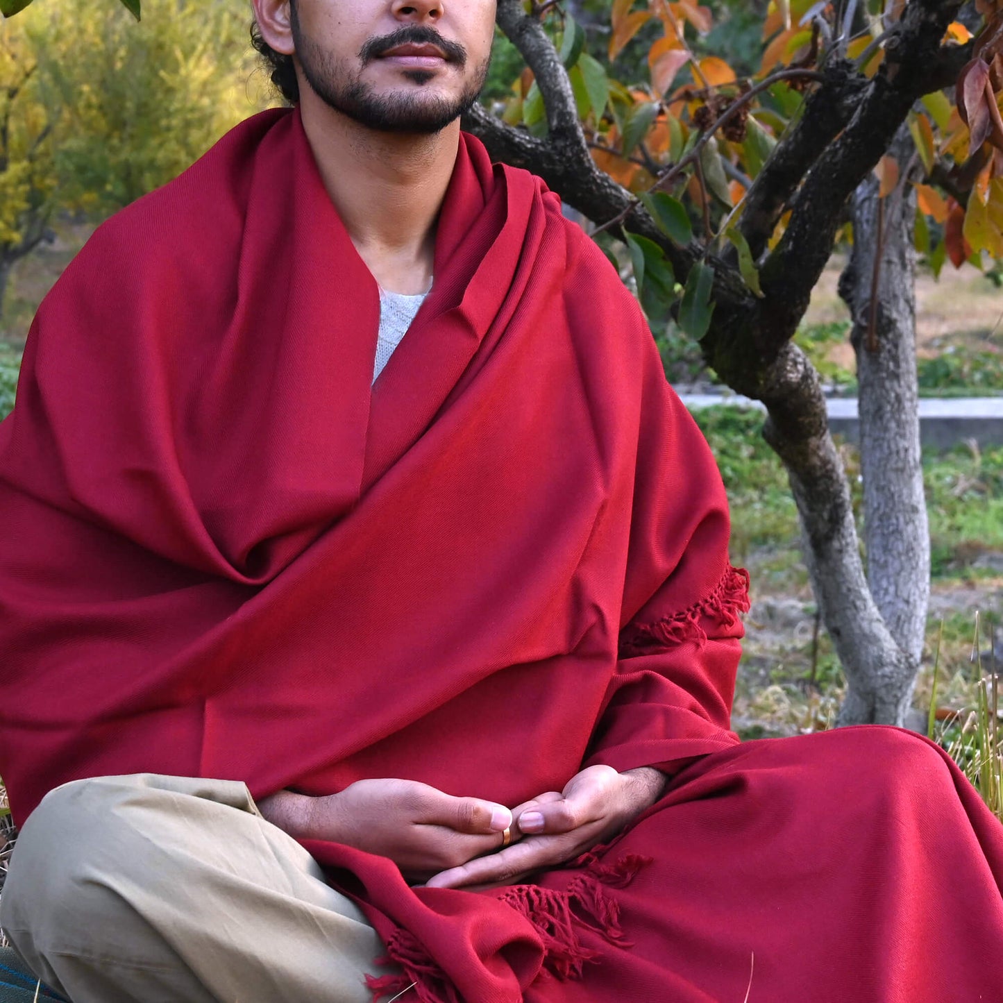 A person seated outdoors wearing a maroon meditation shawl with a fringed edge, sitting next to a tree with autumn leaves.